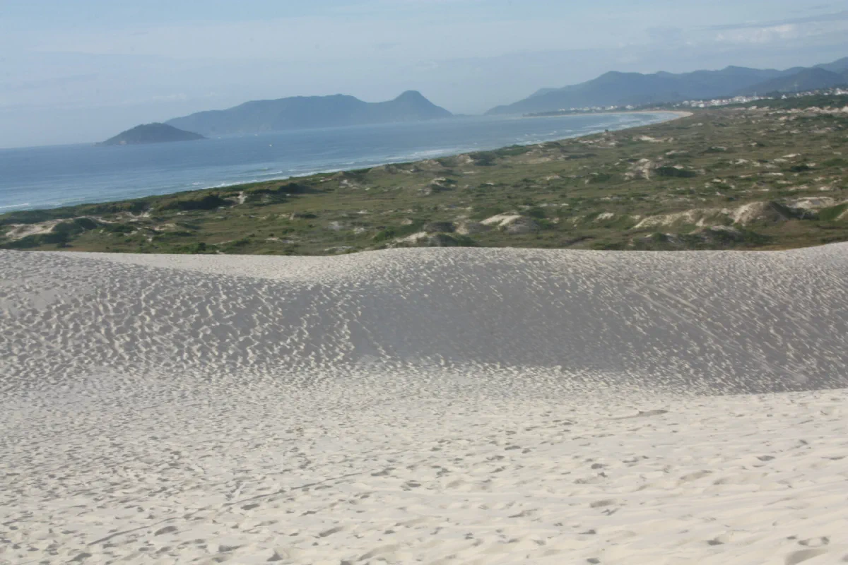 Duna de areia branca em primeiro plano, com a praia e o mar ao fundo.