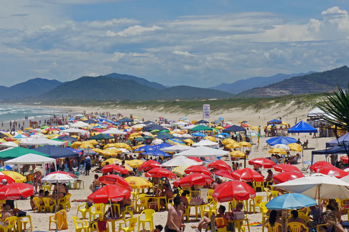 Praia da Joaquina cheia, com guarda-sóis coloridos lado a lado e morros ao fundo.