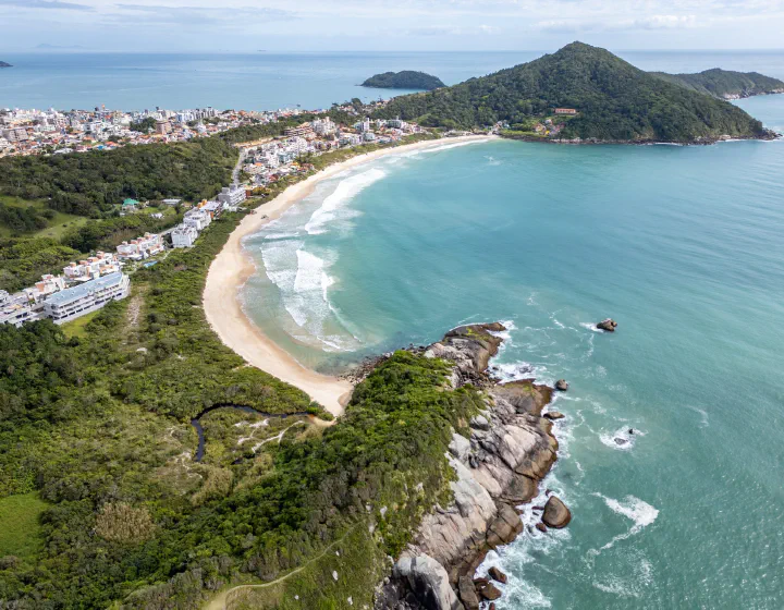 Vista aérea do litoral de Bombinhas (SC), com praia em curva, costão e mar verde-azulado