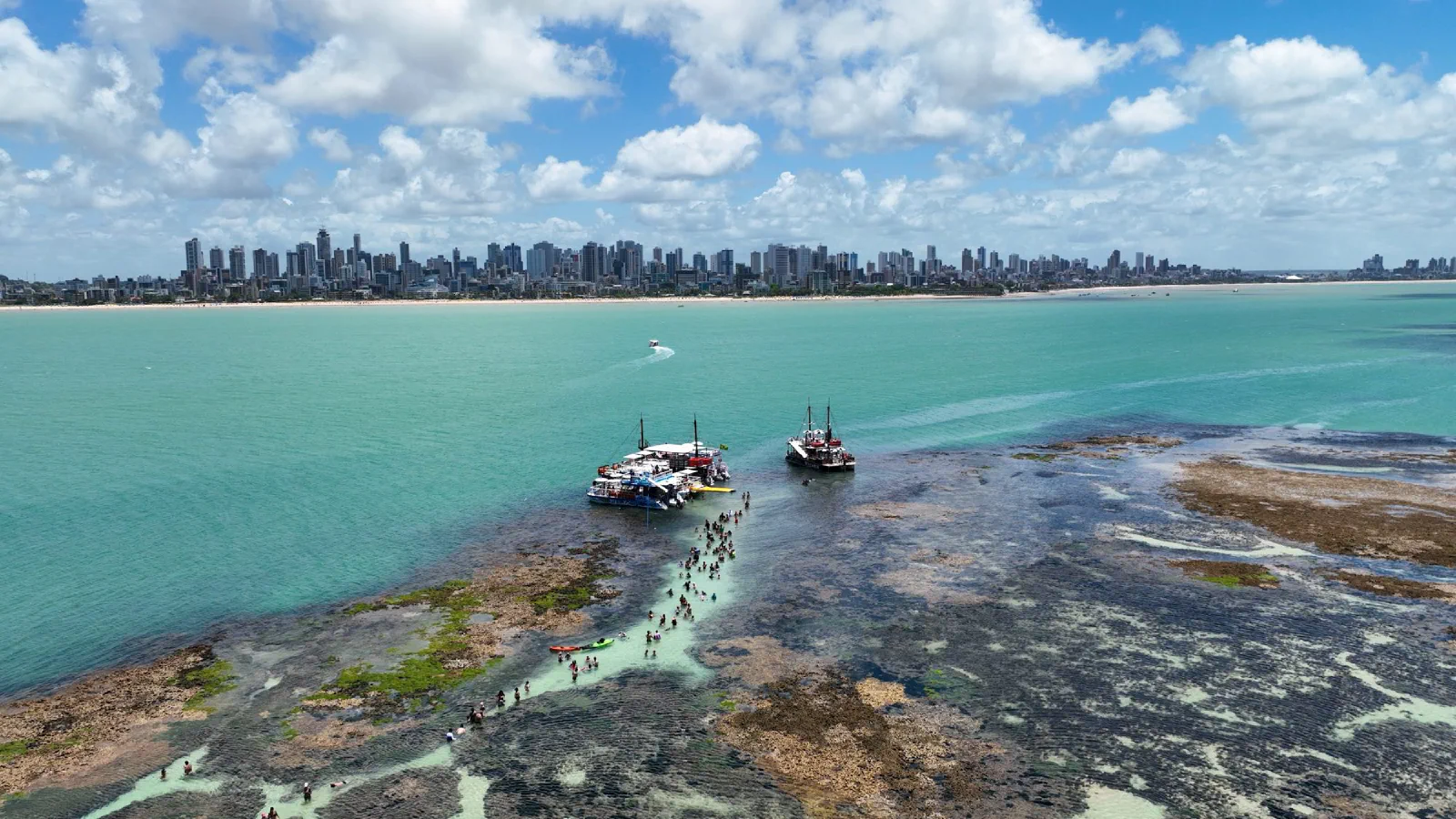 Piscinas naturais do Picãozinho na maré baixa, com barcos e os prédios de João Pessoa (PB) ao fundo