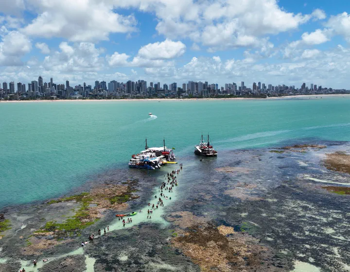 Piscinas naturais do Picãozinho na maré baixa, com barcos e os prédios de João Pessoa (PB) ao fundo