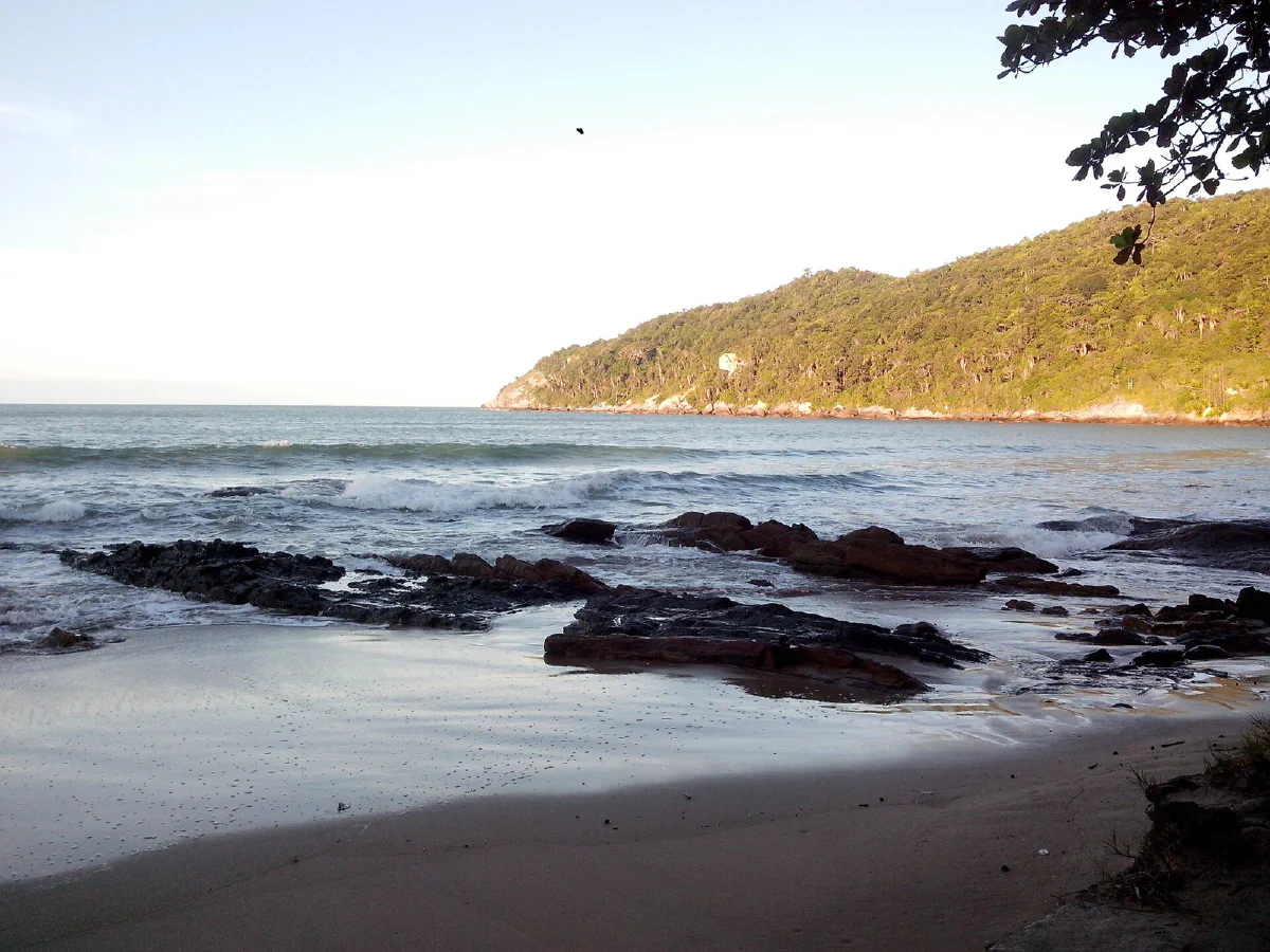 Pedras junto à areia da Sepultura, com o mar e um morro coberto de vegetação ao fundo.