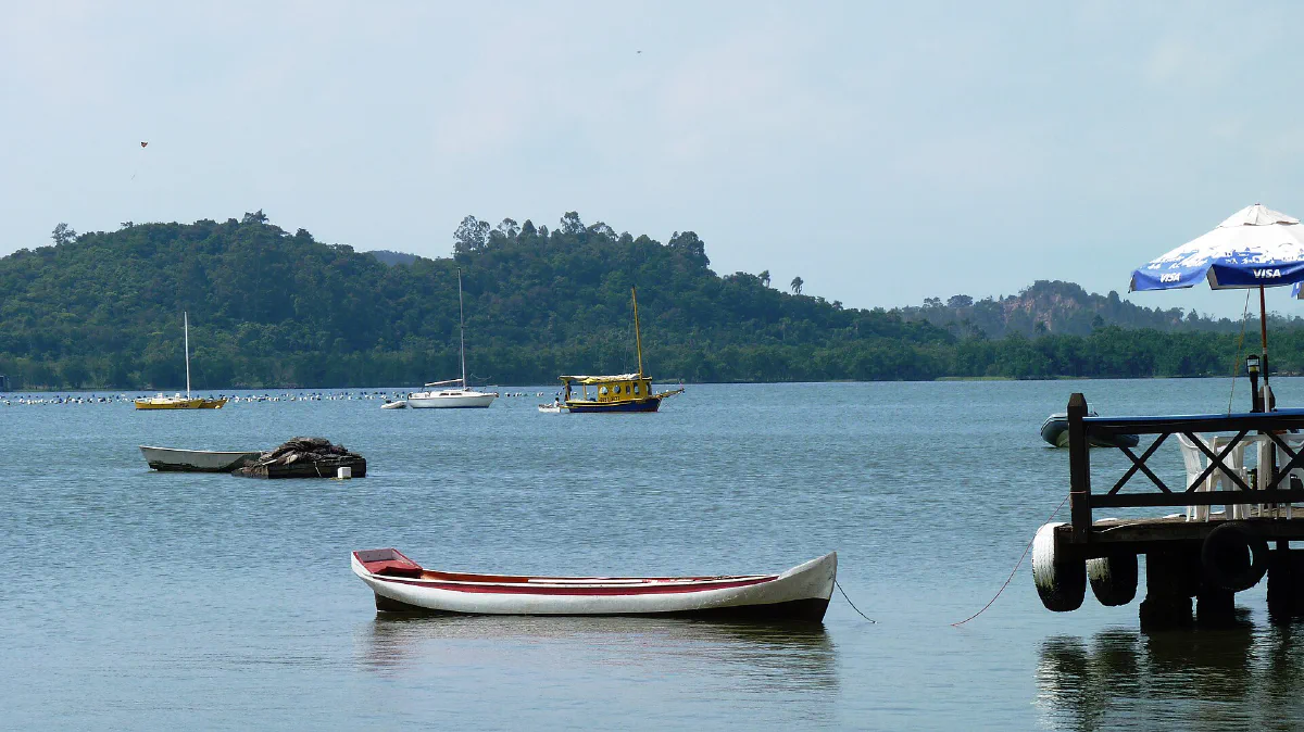Barcos ancorados na baía, com um trapiche de madeira à direita e o morro ao fundo.