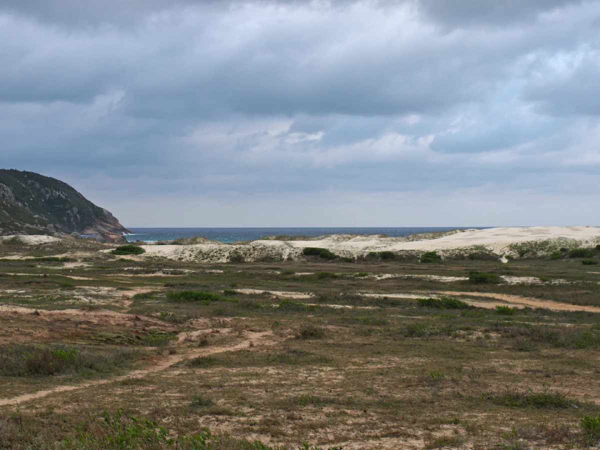 Campo de dunas com o mar e um morro ao fundo.