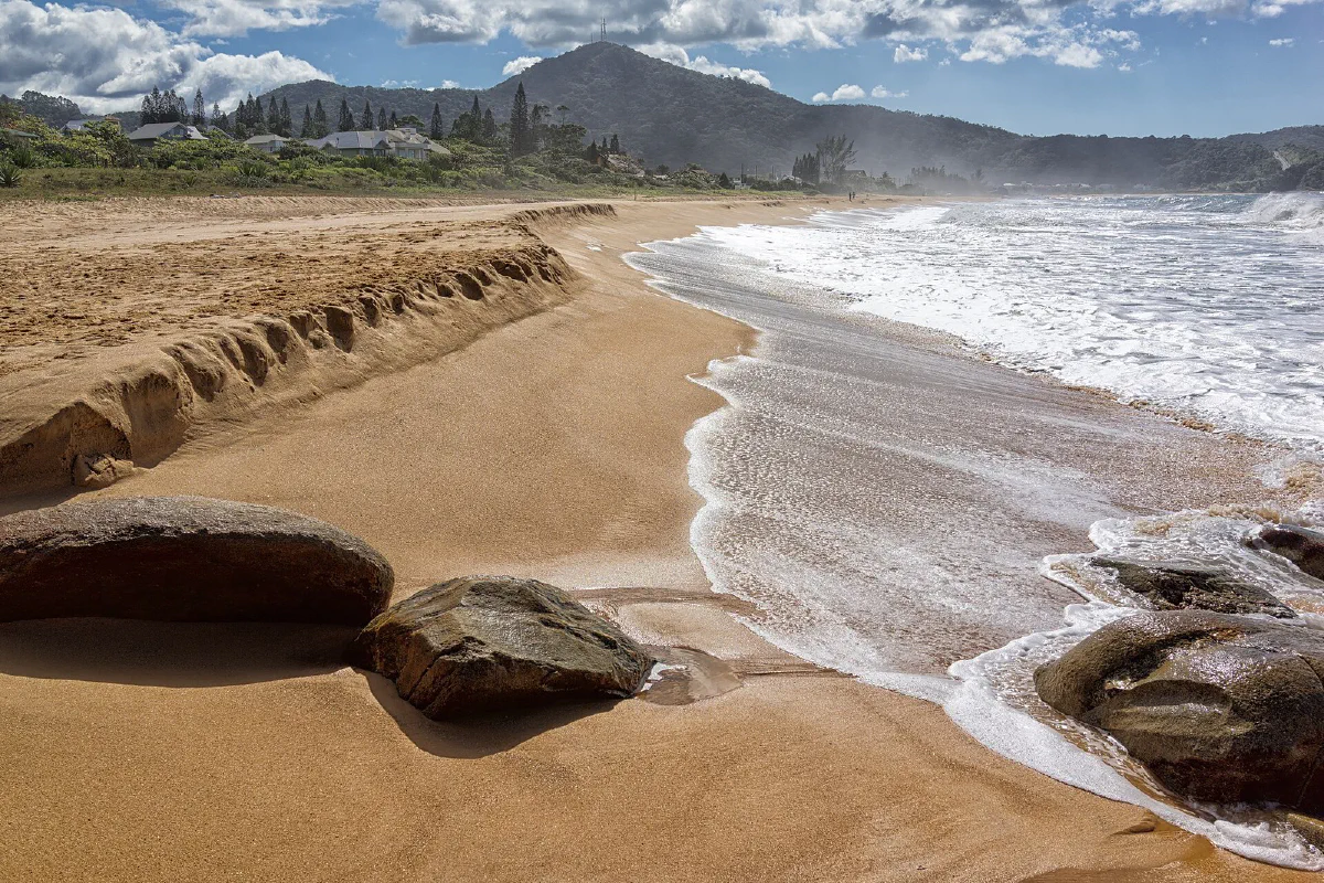 Ondas na Praia do Estaleiro, com pedras em primeiro plano e casas junto à vegetação.