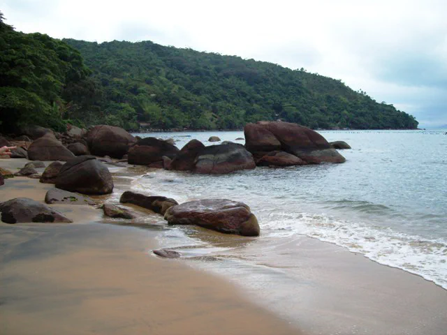 Pedras na beira do mar na Praia da Lagoinha, com morro coberto de mata ao fundo.