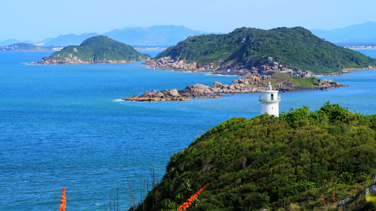 Farol branco num morro verde, com o mar e ilhas ao fundo.