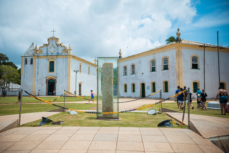 Construções antigas de cor amarela e branco com um monumento ao meio protegido por vidro no Centro Histórico de Porto Seguro na Bahia 