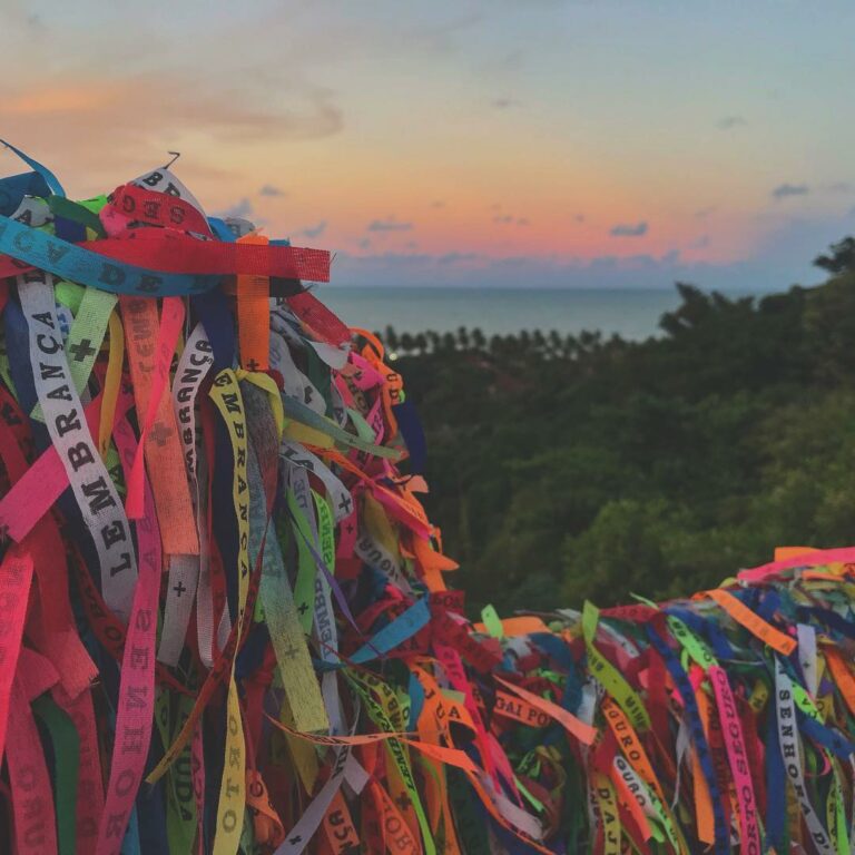 Várias fitinhas do Senhor do Bonfim amarradas em frente a um pôr do sol com uma vegetação do lado no Largo da Ajuda em Arraial d’Ajuda na Bahia